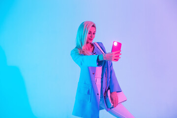 Glamorous young girl in fashionable bright clothes with a stylish handbag makes a selfie photo on a smartphone in the studio on a colored neon light
