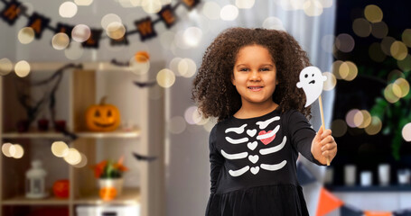 halloween, holiday and childhood concept - smiling african american girl in black costume dress with skeleton bones and ghost over decorated home room and lights background