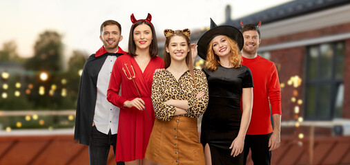 friendship, holiday and people concept - group of happy smiling friends in halloween costumes of vampire, devil, witch and leopard over roof top party background