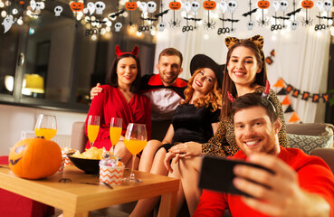 friendship, holiday and people concept - group of happy smiling friends in halloween costumes of vampire, devil, witch and cheetah taking selfie by smartphone at home party at night