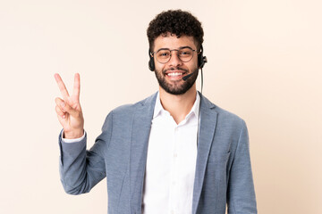 Telemarketer Moroccan man working with a headset isolated on beige background smiling and showing victory sign