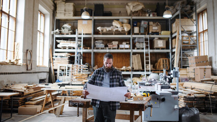 Portrait of mature male carpenter looking at blueprints plans in carpentery workshop. Small business concept.