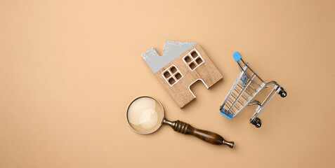 model of a wooden house, a miniature shopping cart and a magnifying glass on a light brown background. Home search concept for rent, purchase, mortgage