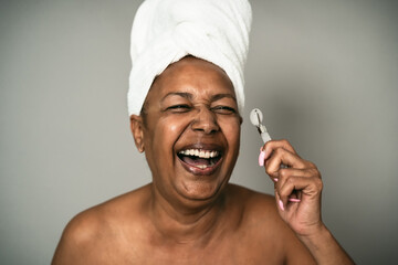 Happy Afro senior woman using jade roller during skin care spa day - People wellness lifestyle...
