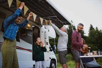 Multi-generation family preparing party by car outdoors in campsite, caravan holiday trip.