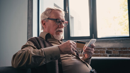 pensioner in eyeglasses using smartphone and holding credit card at home