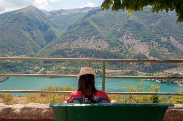 Back view of woman with hat sitting on mountain in front of lake.