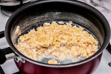 on the stove, a healthy and dietary oatmeal porridge with milk is prepared in a burgundy non-stick...