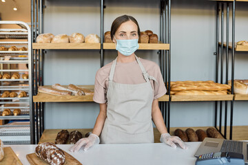Portrait of young smiling female clerk in mask standing by counter against display with bakery assortment