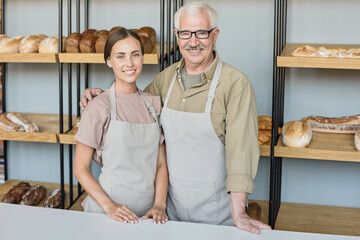 Portrait of happy senior baker in apron embracing young daughter while working with her in own...