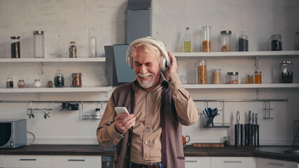 happy and senior man in wireless headphones listening music and holding smartphone in kitchen