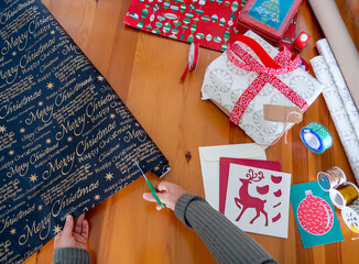Fototapeta premium Woman hands cutting black Christmas wrapping paper. Work station for Xmas gifts decoration with ribbon, string and holiday cards. Top view of Christmas season flat lay with Christmas pattern