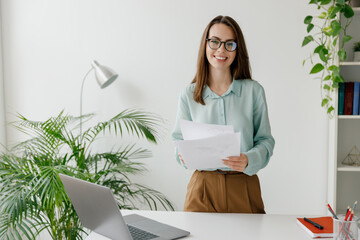 Young confident employee business woman in blue shirt hold paper account documents work stand at...