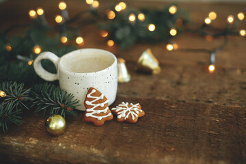 Christmas gingerbread cookies, coffee cup, fir branches on background of warm lights bokeh on rustic wooden table. Moody atmospheric image. Winter countryside hygge. Seasons greeting