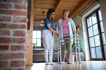 Healthcare worker or caregiver visiting senior woman indoors at home, helping her to walk.