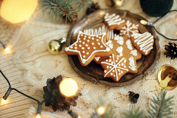 Homemade christmas gingerbread cookies on plate, fir branches, ornaments and warm lights on cozy knitted background. Atmospheric festive time and hygge home. Happy Holidays. Hello winter