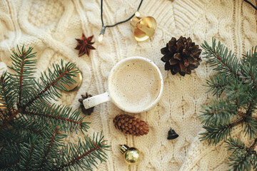 Obraz na płótnie Canvas Warm coffee in stylish cup, anise star, fir branches, ornaments, pine cones and warm lights on cozy knitted background. Top view. Atmospheric christmas time and hygge winter home