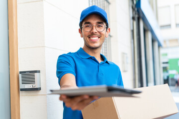 Young delivery man at outdoors holding boxes and a tablet with happy expression