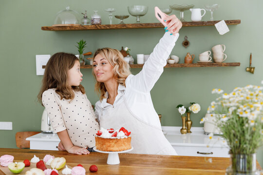 Happy fun chef cook baker mom woman in white shirt work baby girl helper do selfie shot on mobile cell phone at kitchen table home Cooking food process concept Mommy little kid daughter prepare cake