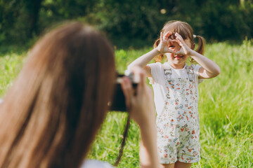 Obraz premium Photographer woman in white clothes take picture shot on photo camera of child baby girl 5-6 years old show heart-shape sign gesture Mommy rest with little kid daughter outdoor. Love family concept