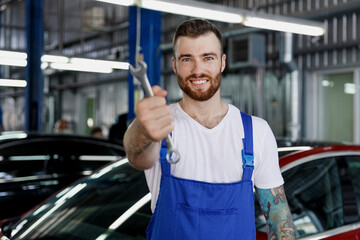 Smiling happy young male professional technician car mechanic repairman man 20s in denim blue overalls white t-shirt hold give wrench key tool work in light modern vehicle repair shop workshop indoor