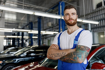 Strong minded young technician car mechanic man in denim blue overalls white t-shirt look aside hold hands crossed folded work in light modern vehicle repair shop workshop indoor Tattoo translate fun