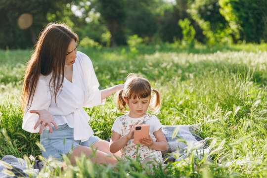Indignant young woman wear white clothes have fun picnic sit on plaid grass with child baby girl 5-6 years old playing mobile cell phone Mommy playing rest with little kid daughter outdoor together