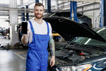 Young professional technician car mechanic man in overalls white t-shirt hold mobile cell phone with blank screen area fix problem with raised hood bonnet work in vehicle repair shop workshop indoor