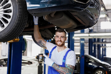 Strong smiling happy young male professional technician mechanic man in denim blue overalls white t-shirt point finger on car lift check technical condition work in vehicle repair shop workshop inside