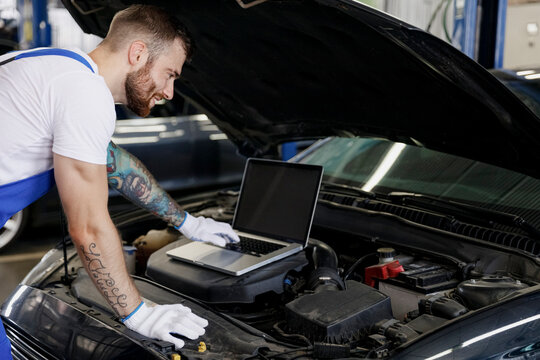 Young repairman professional technician car mechanic man in blue overalls t-shirt use laptop pc computer make diagnostics check fix problem with raised hood work in vehicle repair shop workshop inside