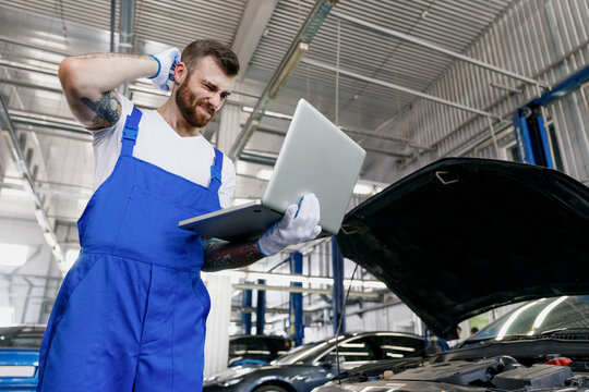 Lower underside young professional technician car mechanic man in overalls t-shirt use hold laptop pc computer make diagnostics fix problem with raised hood work in vehicle repair shop workshop inside