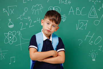 Young shrewd male kid school boy 5-6 years old in t-shirt backpack hold hands crossed isolated on green wall chalk blackboard background studio. Childhood children kids education lifestyle concept