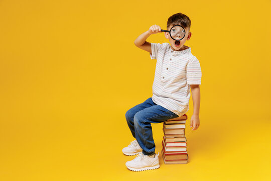 Side view young school boy 5-6 year old in casual clothes sit on pile of books looking through magnifier isolated on plain yellow background studio Childhood children kids education lifestyle concept