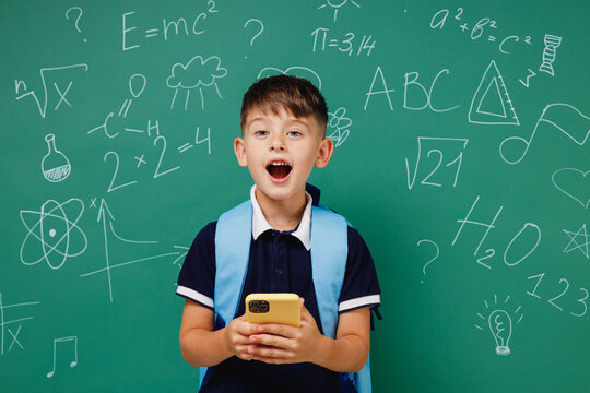 Young amazed fun male kid school boy 5-6 years old in t-shirt backpack using mobile cell phone isolated on green wall chalk blackboard background. Childhood children kids education lifestyle concept