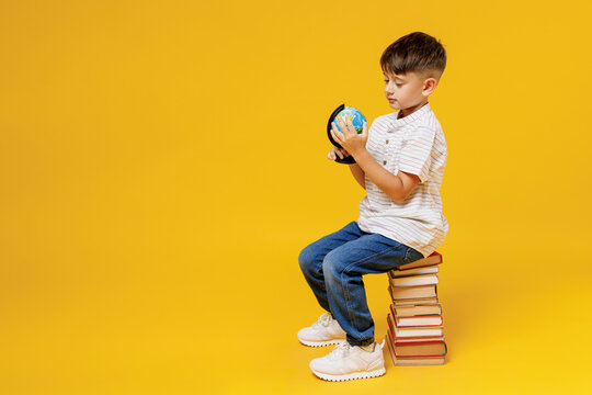 Side view young school boy 5-6 years old in casual clothes sit on pile of books spin Earth world globe isolated on plain yellow background studio Childhood children kids education lifestyle concept.