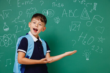 Young male kid school boy 5-6 years old in t-shirt backpack point hand aside on workspace area isolated on green wall chalk blackboard background. Childhood children kids education lifestyle concept.