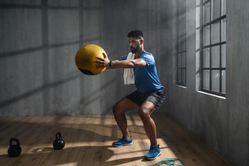 Young African American sportsman standing and lifting a medicine ball indoors, crossfit training concept.