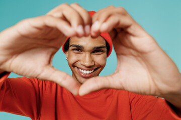 Close up young smiling attractive happy african american man in orange shirt hat show shape heart look through hands heart-shape sign isolated on plain pastel light blue background studio portrait