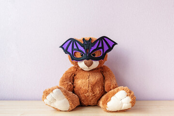 Funny brown teddy bear with purple bat shaped mask sits on wooden table by light pink wall in room....