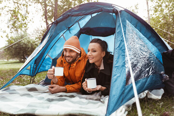 camping, tourism and travel concept - happy couple lying inside tent and drinking tea at campsite