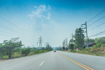 Fototapeta premium An image of a car cruising along an open road with trees and lampposts along the way against a background of sky and clouds.