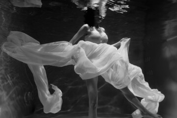 Black and white photo where a beautiful girl in a white dress swims underwater in the pool, and she looks like a siren or a mermaid
