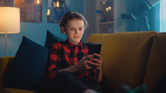 Cute Young Boy Sitting on a Sofa in a Dark Cozy Room and Using a Smartphone at Home. Happy Teenager Browsing Content Online, Chatting with Friends on Social Media, Reading Articles on Internet. © Gorodenkoff