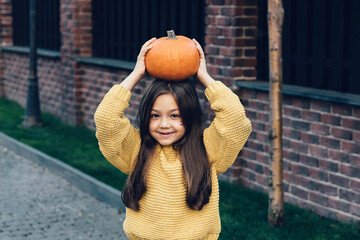Funny child girl in orange pullover for Halloween with pumpkin and on a dark brick background.