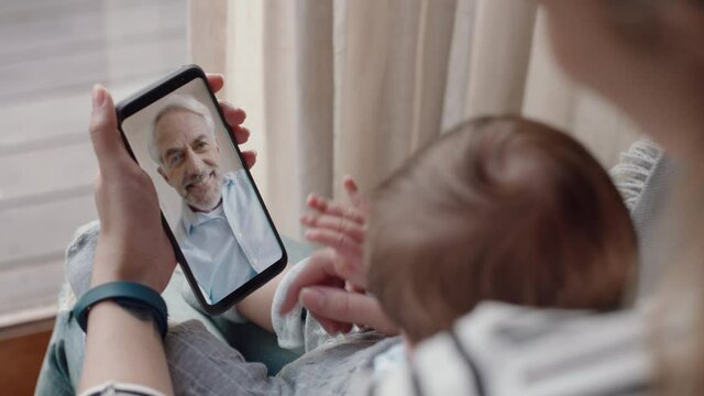 young mother and baby having video chat with grandfather using smartphone waving at newborn infant enjoying family connection