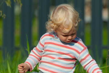 a blond curly-haired boy in a striped sweater with his head down is sitting in the grass on the...