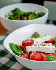 Caprese salad with strawberry, mozzarells, basil and balsamic sauce served in a white bowl over green plaid background.
