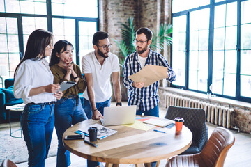 Group of diverse male and female coworkers checking received paper letter with news about startup investment, skilled hipster guys using modern technology during collaborative meeting for studying