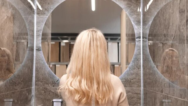 A blonde woman stands with her back to the camera and turns on the illumination of the mirror that hangs in the bathroom