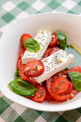 Caprese salad with strawberry, mozzarells, basil and balsamic sauce served in a white bowl over green plaid background.